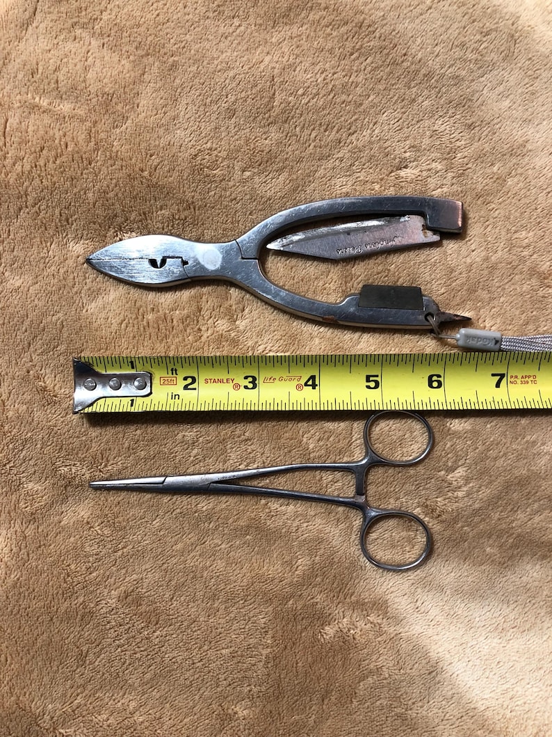May include: Two surgical tools, a pair of pliers with a small knife and a pair of hemostats, are laid out on a brown fuzzy surface. A yellow measuring tape is placed between the tools. The tape reads 2, 3, 4, 5, 6, and 7 inches.