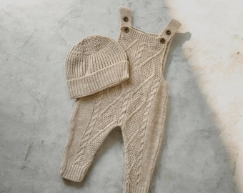 May include: A beige knitted baby outfit set, including a beanie and overalls. The overalls feature a cable knit design and button closures on the straps. The set is made of a soft, textured material, perfect for a newborn.