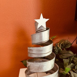 May include: A silver metal Christmas tree sculpture with a white star on top. The tree is made of a spiral of metal and is sitting on a white surface.