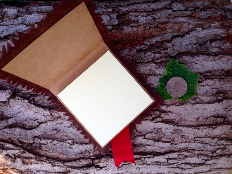 May include: A small, brown, leather-bound journal with a red ribbon bookmark. The journal is open and lying on a tree bark background. A silver coin is visible on a green leaf to the right of the journal.