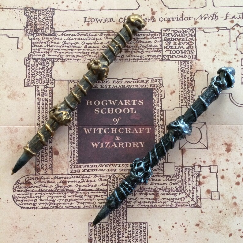 May include: Two decorative wands, one with a black and silver finish and the other with a gold and brown finish. Both wands have intricate details and are displayed on a map of Hogwarts School of Witchcraft and Wizardry.