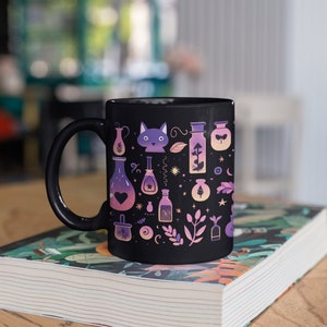 May include: Black ceramic mug with a whimsical design featuring a cat, potion bottles, and other magical elements in shades of purple, pink, and blue.