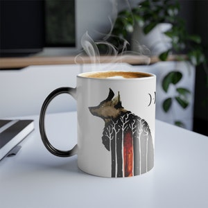 May include: White ceramic coffee mug with a black handle. The mug features a black and white illustration of a wolf with a forest inside of it. The wolf is silhouetted against a red and orange background.