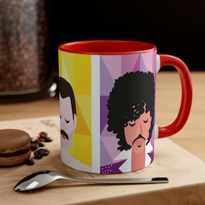 May include: A red and white ceramic coffee mug with a geometric design featuring illustrations of Freddie Mercury and Prince. The mug has a red handle.