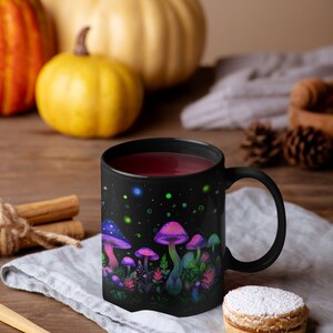 May include: Black ceramic mug with a colorful illustration of glowing mushrooms and foliage. The mug is filled with a dark red liquid and is sitting on a gray linen napkin.