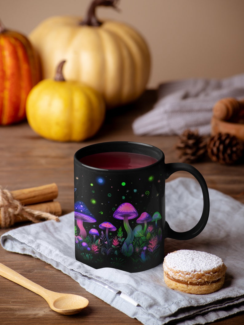 May include: Black ceramic mug with a colorful illustration of glowing mushrooms and foliage. The mug is filled with a dark red liquid and is sitting on a gray linen napkin.