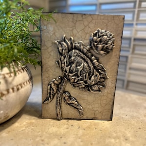 May include: A rectangular, off-white decorative tile with a raised relief of a sunflower. The sunflower is detailed in shades of gray and black, with a cracked, aged appearance. The tile is likely for home decor.