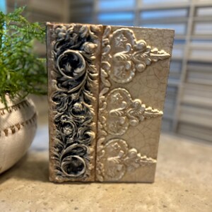 May include: A decorative, rectangular box with an antique book design. The box features ornate, raised floral and scrollwork patterns in cream and black. The box has a cracked, aged appearance, suggesting a vintage aesthetic.