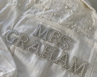 Könnte beinhalten: Cremefarbene Jeansjacke mit verstreuten Perlen und dem Namen "MRS GRAHAM" aus Perlen- und Strassbuchstaben. Die Jacke hat einen klassischen Kragen und geknöpfte Manschetten.