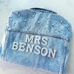 Puede incluir: Una chaqueta vaquera azul claro con un parche de "MRS BENSON" adornado con perlas blancas en la espalda. La chaqueta está decorada con pequeños diamantes de imitación.