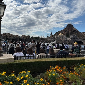 May include: A large crowd of people gathered in front of a large, gray, stone building with a volcano in the background. The building has a large, golden dome and a tall, white tower. The people are standing and sitting on a stone walkway. There are yellow flowers in the foreground.