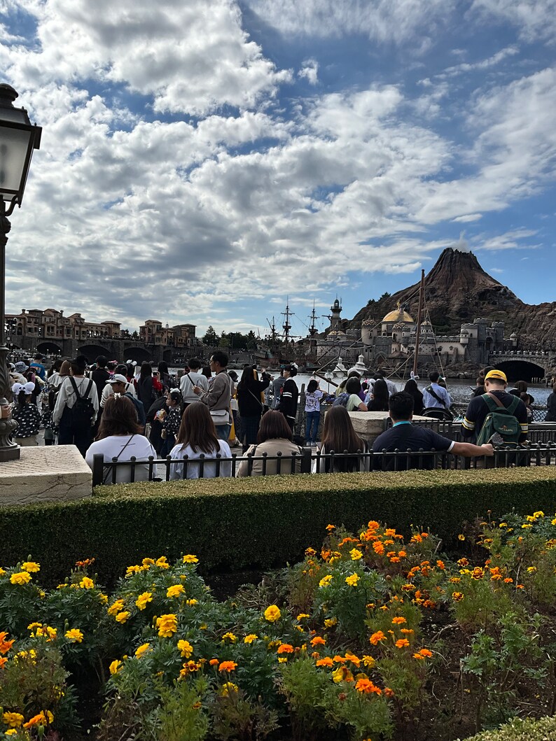 May include: A large crowd of people gathered in front of a large, gray, stone building with a volcano in the background. The building has a large, golden dome and a tall, white tower. The people are standing and sitting on a stone walkway. There are yellow flowers in the foreground.