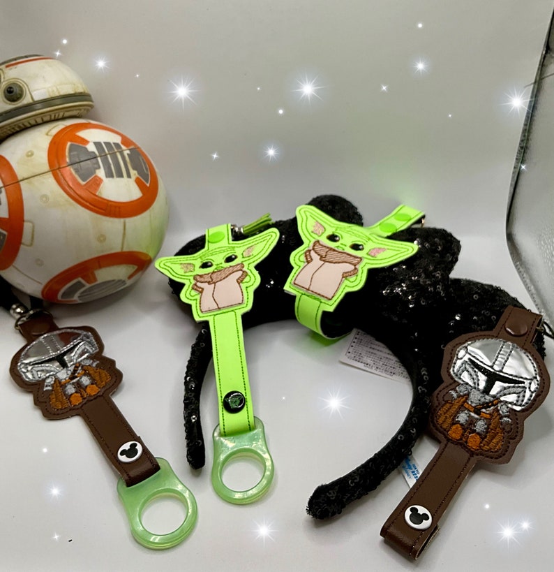 K&ouml;nnte beinhalten: Eine Sammlung von Star Wars-Accessoires, darunter ein BB-8-Droide, Charakter-Schl&uuml;sselanh&auml;nger und ein Paar schwarze, paillettenbesetzte M&auml;useohren mit Grogu und neongr&uuml;nen Akzenten. Der Mandalorianer-Schl&uuml;sselanh&auml;nger ist braun und silber.