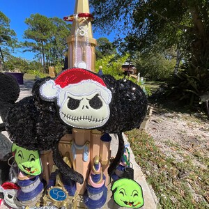 May include: A pair of black and white Mickey Mouse ears with a black sequin fabric. The ears are decorated with a green and black character with a toothy grin and a red and white character with a black and white face and a Santa hat. The ears are in front of a gold and white castle.