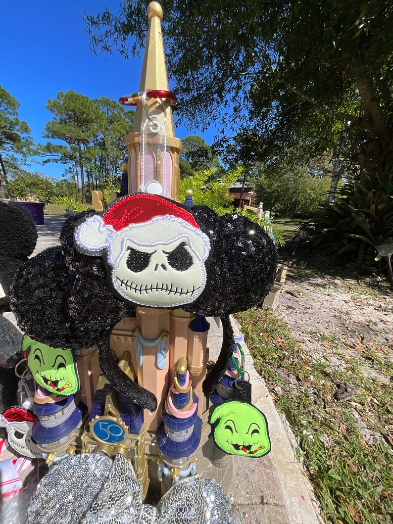 May include: A pair of black and white Mickey Mouse ears with a black sequin fabric. The ears are decorated with a green and black character with a toothy grin and a red and white character with a black and white face and a Santa hat. The ears are in front of a gold and white castle.