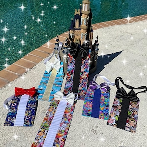 May include: Six different bow tie keychains with various Disney character designs. The keychains are displayed on a gray surface with a miniature castle in the background.