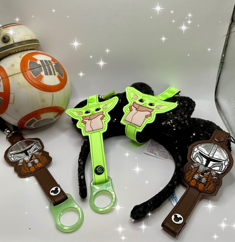 K&ouml;nnte beinhalten: Eine Auswahl an Star Wars-Accessoires. Enth&auml;lt zwei gr&uuml;ne und beige Grogu (Baby Yoda) Ohrhalter, zwei braune Mandalorianer Ohrhalter und ein BB-8-Droiden-Spielzeug. Die Ohrhalter haben ein Mickey Mouse-Symbol.