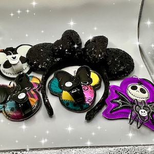 Może przedstawiać: Pięć akcesori&oacute;w do włos&oacute;w inspirowanych postaciami Disneya w stylu Halloween. Akcesoria są wykonane z tkaniny i cekin&oacute;w. Akcesoria przedstawiają uszy Myszki Miki, Jacka Skellingtona i fioletową postać z nietoperzowymi skrzydłami.