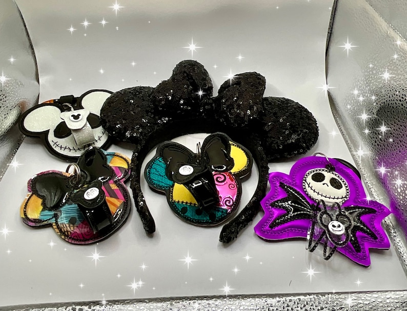 Może przedstawiać: Pięć akcesori&oacute;w do włos&oacute;w inspirowanych postaciami Disneya w stylu Halloween. Akcesoria są wykonane z tkaniny i cekin&oacute;w. Akcesoria przedstawiają uszy Myszki Miki, Jacka Skellingtona i fioletową postać z nietoperzowymi skrzydłami.