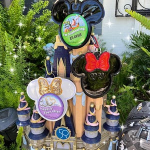 May include: Three black and white Mickey Mouse ears with a bow, a gold glitter bow, and a green and white button that says "I'm Celebrating: Eloise" on a miniature castle with a pink and blue roof. The castle has a sign that says "Happily Ever After" and a number 50 on it.