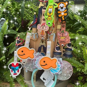 May include: A set of colorful Disney character keychains featuring Goofy, Tigger, Mickey Mouse, and a fish. The keychains are attached to a light blue headband with a sparkly silver ear and a light blue bow. The keychains are displayed on a miniature castle.