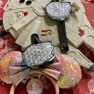May include: A pair of pink and iridescent sequined Minnie Mouse ears with a black and silver Millennium Falcon charm attached. The charm is on a black leather strap with a silver buckle. The ears are in front of a white Millennium Falcon toy.