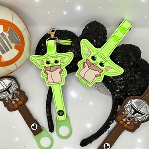 K&ouml;nnte beinhalten: Gr&uuml;ne und braune Baby Yoda und Mandalorianer Accessoires. Die Baby Yoda Accessoires sind hellgr&uuml;n mit beige und rosa Akzenten. Die Mandalorianer Accessoires sind braun mit silbernen Akzenten. Die Accessoires haben einen goldenen Verschluss.