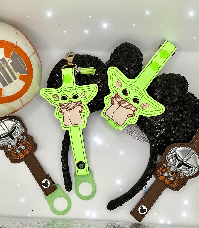 K&ouml;nnte beinhalten: Gr&uuml;ne und braune Baby Yoda und Mandalorianer Accessoires. Die Baby Yoda Accessoires sind hellgr&uuml;n mit beige und rosa Akzenten. Die Mandalorianer Accessoires sind braun mit silbernen Akzenten. Die Accessoires haben einen goldenen Verschluss.