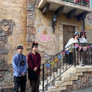 May include: Four people stand in front of a building with a stone facade. The building has a balcony with flowers and a sign that says "La Papillon." There is a large pink number 40 on the wall with the words "Dream Go Rounds." The people are wearing casual clothing and are smiling.
