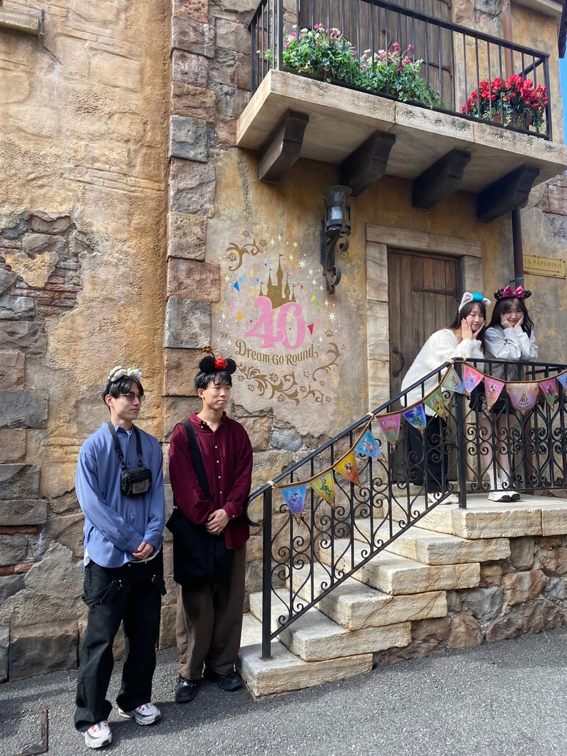 May include: Four people stand in front of a building with a stone facade. The building has a balcony with flowers and a sign that says "La Papillon." There is a large pink number 40 on the wall with the words "Dream Go Rounds." The people are wearing casual clothing and are smiling.
