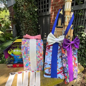 May include: A collection of Disney-themed items including a Toy Story alien figure in a box, three fabric gift bags with large bows, and five white fabric bookmarks with small character icons at the top, all arranged on a stone surface.