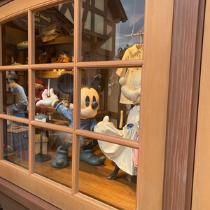 Puede incluir: Un escaparate con una figura de Mickey Mouse que lleva una camisa azul y sostiene una aguja e hilo. La figura est&aacute; de pie junto a un maniqu&iacute; blanco que lleva un vestido blanco con ribetes azules. El escaparate est&aacute; enmarcado en madera con un marco de madera marr&oacute;n.