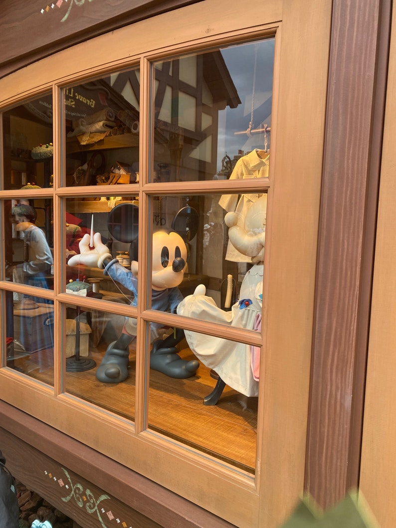 Puede incluir: Un escaparate con una figura de Mickey Mouse que lleva una camisa azul y sostiene una aguja e hilo. La figura est&aacute; de pie junto a un maniqu&iacute; blanco que lleva un vestido blanco con ribetes azules. El escaparate est&aacute; enmarcado en madera con un marco de madera marr&oacute;n.
