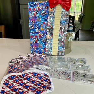 May include: A red and white striped fabric with blue Mickey Mouse ears, a blue and white gift bag with a red bow, and several clear plastic bags with glitter inside.