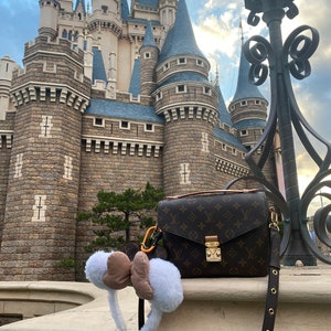 May include: A brown leather handbag with a monogram pattern and a gold clasp. The bag is hanging from a lamppost in front of a large stone castle.