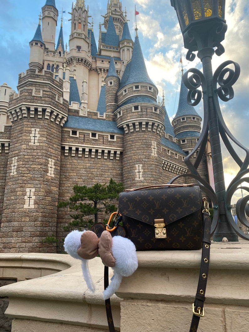May include: A brown leather handbag with a monogram pattern and a gold clasp. The bag is hanging from a lamppost in front of a large stone castle.
