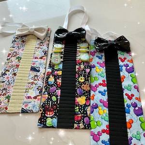 May include: Three fabric bow tie holders with different Disney-themed prints. The bow ties are black with a white satin ribbon. The prints include a colorful Mickey Mouse balloon design, a black and white print with Mickey Mouse and other Disney characters, and a white print with a gold stripe and various Disney characters.