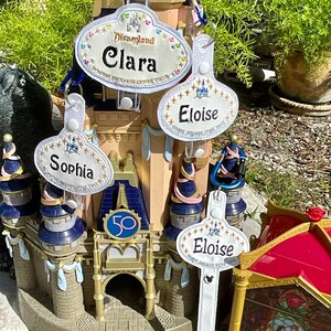 May include: White oval-shaped name tags with gold glitter and embroidery. The tags feature the Disney castle logo and the names "Clara", "Eloise", and "Sophia".