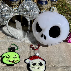 May include: A set of three Nightmare Before Christmas themed Disney merchandise items. A silver sequined Minnie Mouse headband, a white Jack Skellington light-up head, and two keychains featuring Jack Skellington and Oogie Boogie.