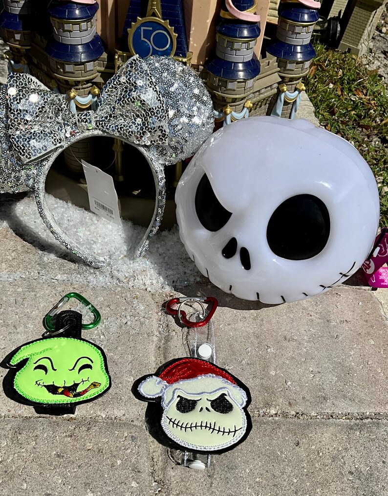 May include: A set of three Nightmare Before Christmas themed Disney merchandise items. A silver sequined Minnie Mouse headband, a white Jack Skellington light-up head, and two keychains featuring Jack Skellington and Oogie Boogie.