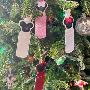 May include: Four different keychains with a Mickey Mouse design. The keychains are made of fabric and have different colors and patterns. The keychains are hanging on a Christmas tree.