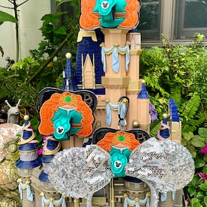 May include: Three Disney character-themed hair bows with a silver glitter bow, a red-haired princess with a green dress, and a red-haired princess with a blue dress. The bows are displayed on a miniature castle.