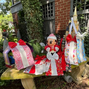 May include: A collection of fabric gift bags and table runners featuring holiday-themed patterns, including red and white swirls, gingerbread men, and character prints. Each item is accented with large satin bows and white ribbon detailing.