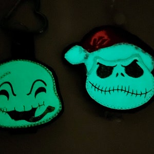 May include: Two glow-in-the-dark felt ornaments featuring characters from the Nightmare Before Christmas. One ornament is green and features a smiling ghost with a stitched mouth. The other ornament is white and features Jack Skellington wearing a Santa hat.