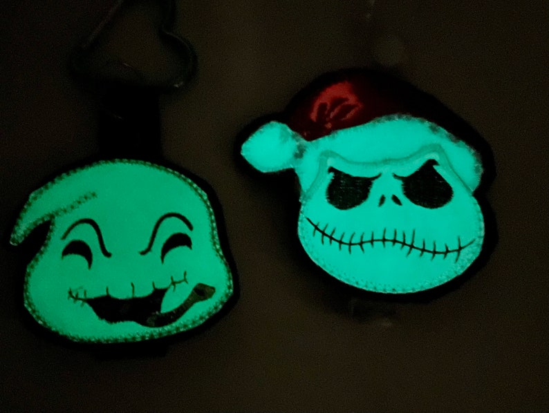 May include: Two glow-in-the-dark felt ornaments featuring characters from the Nightmare Before Christmas. One ornament is green and features a smiling ghost with a stitched mouth. The other ornament is white and features Jack Skellington wearing a Santa hat.