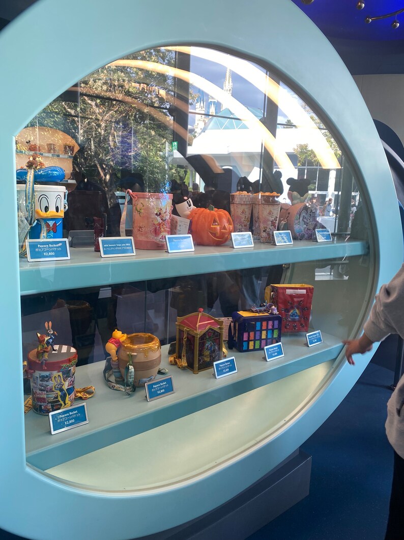 Puede incluir: Una vitrina de vidrio con una variedad de cubos de palomitas de ma&iacute;z coloridos con personajes de Disney. Los cubos est&aacute;n dispuestos en dos estantes. El estante superior presenta un cubo de calabaza de Mickey Mouse, un cubo de Donald Duck y un cubo de Winnie the Pooh. El estante inferior presenta una variedad de otros cubos, incluyendo un cubo de Cenicienta, un cubo de Peter Pan y un cubo de Mickey Mouse. La vitrina est&aacute; ubicada en un parque de Disney.
