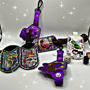 May include: Five different purple and white fabric keychains with various designs. The designs include a frog, a Hogwarts house crest, a Gryffindor crest, a Harry Potter character, and a Hogwarts house crest.