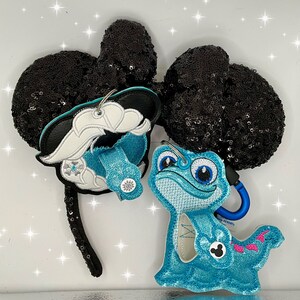 Puede incluir: Orejas de Minnie Mouse de lentejuelas negras con un amuleto de personaje Olaf azul y blanco y un amuleto de personaje azul y turquesa de un lagarto azul con cola rosa.