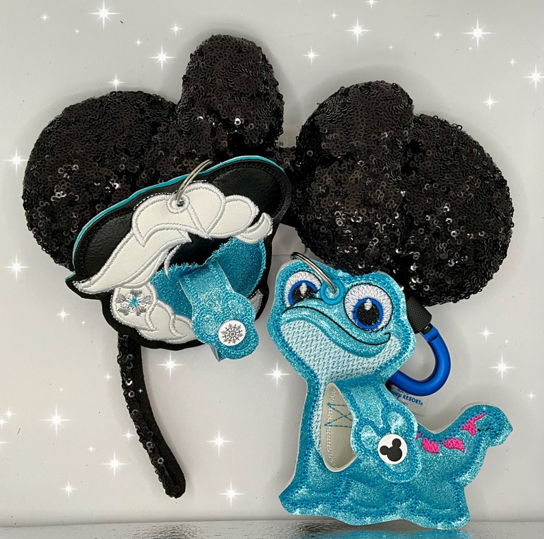 Puede incluir: Orejas de Minnie Mouse de lentejuelas negras con un amuleto de personaje Olaf azul y blanco y un amuleto de personaje azul y turquesa de un lagarto azul con cola rosa.