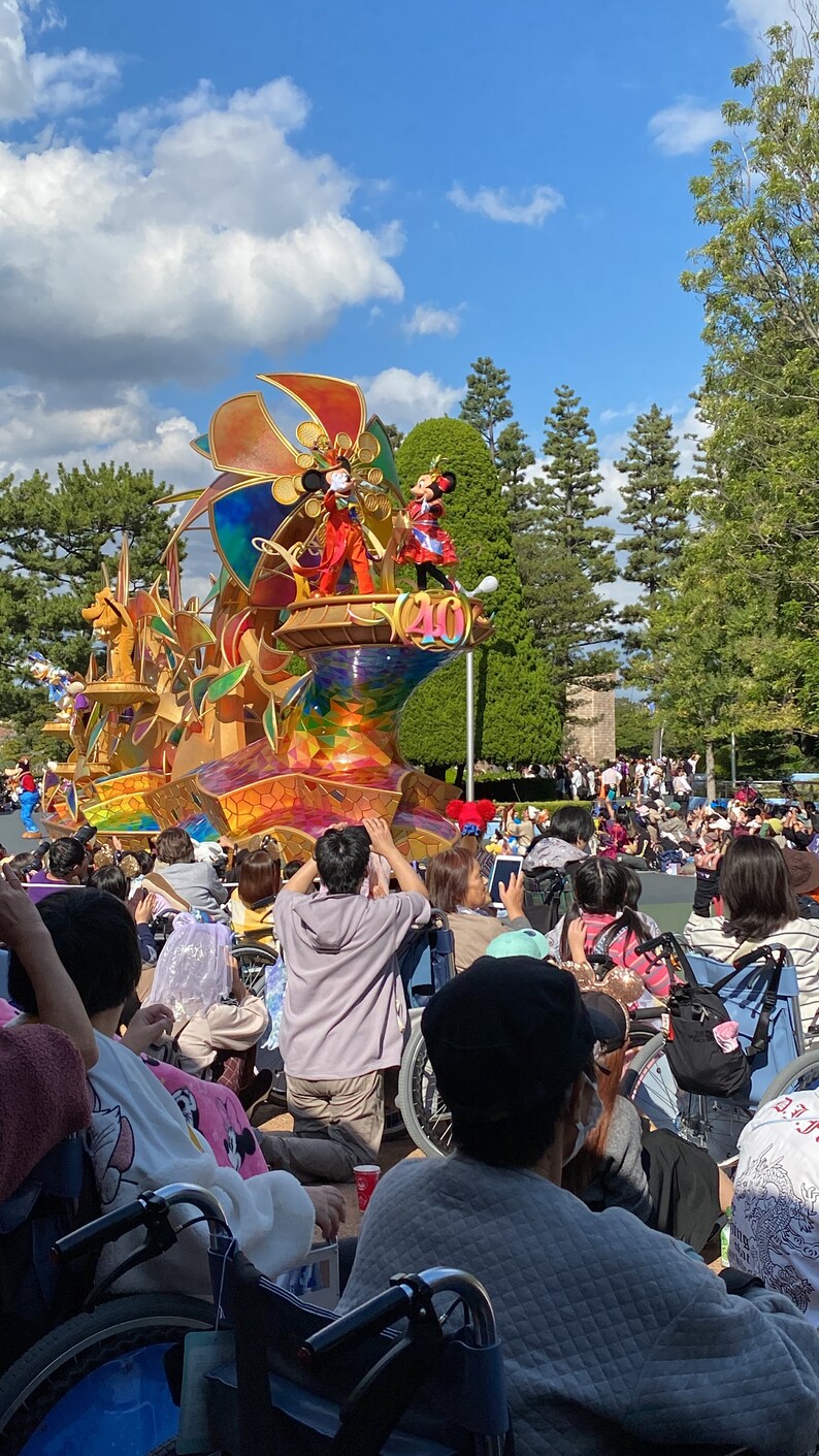 Puede incluir: Un carro aleg&oacute;rico colorido y elaborado con un gran dise&ntilde;o de molino de viento dorado. Mickey Mouse y Minnie Mouse est&aacute;n en el carro, saludando a la multitud. Las personas est&aacute;n sentadas en sillas de ruedas, viendo el desfile.
